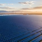 Aerial oblique view of a vast solar farm at golden hour, blue photovoltaic panels stretching toward distant transmission towers and battery storage units with low mountains under soft clouds.