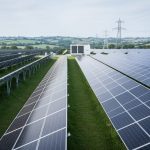 Wide view of a UK utility-scale solar farm with tilted solar panel trackers, distant grid substation structures, and a battery storage unit under an overcast sky.